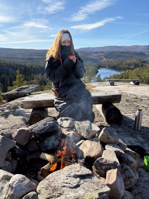 en person sitter ved et bål omgitt av steiner, iført utekos mikrofiber™-plagg, med fjell og skog i bakgrunnen
