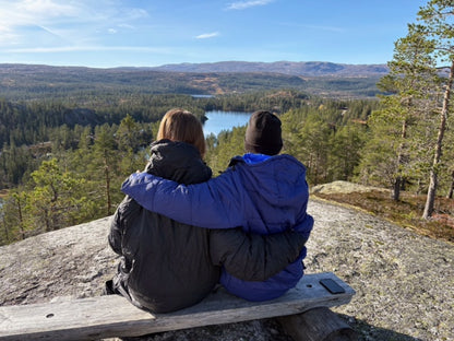 to personer sitter på en trebenk med utsikt over fjell og innsjø, iført varme utekos mikrofiber™-plagg for friluftsliv og tur